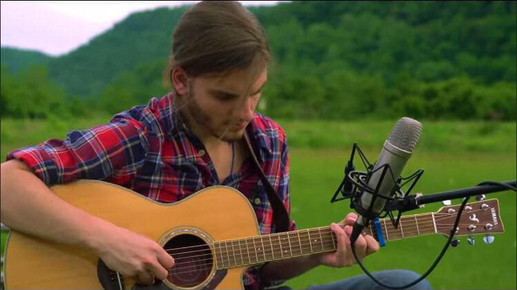 Sam Epllin wearing a red and blue plaid shirt with a guitar and a microphone